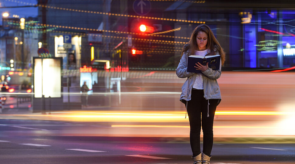 Eine Frau steht abends an einer Straße und liest in einer Bibel. Sie steht ruhig auf der Stelle. Um sie herum fahren Autos vorbei und hinterlassen Lichtstreifen auf dem Bild. Symbolbild für Ruhe und Gebet. 160324-93-000070.jpg