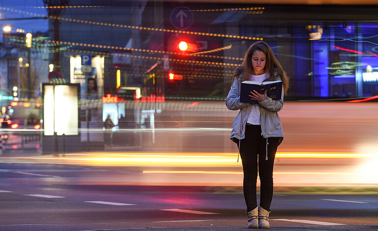 Eine Frau steht abends an einer Straße und liest in einer Bibel. Sie steht ruhig auf der Stelle. Um sie herum fahren Autos vorbei und hinterlassen Lichtstreifen auf dem Bild. Symbolbild für Ruhe und Gebet.