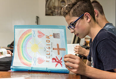 Ein Schüler blättert in seiner Arbeitsmappe beim katholischen Religionsunterricht in einer achten Klasse. Auf das Deckblatt hat er christliche Symbole gezeichnet: ein Christusmonogramm, ein Kreuz, eine Kerze, Fischsymbole, sowie Regenbogen, Sonne und Herzen.