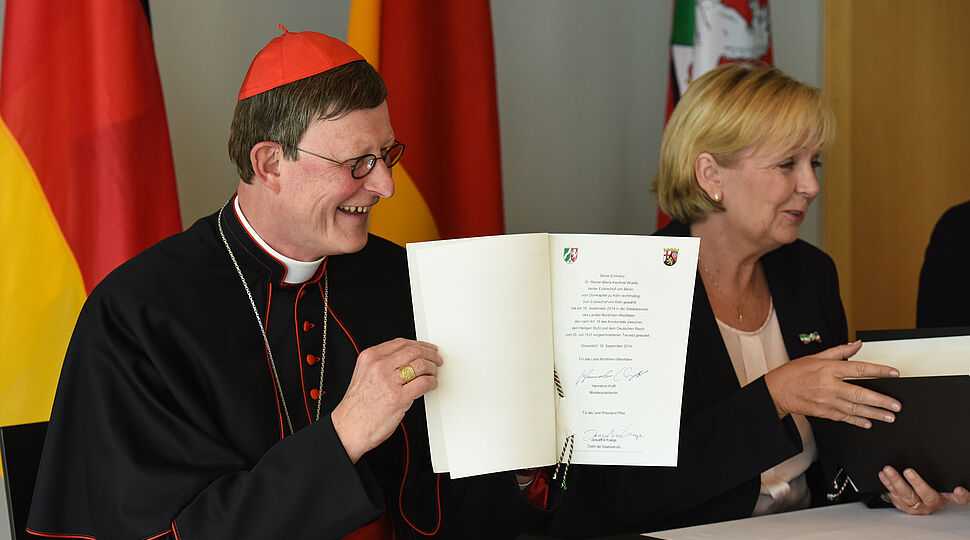 Beide sitzen an einem Tisch und Kardinal Woelki hält ein Dokument in die Höhe. Kirche-Staat_140918-93-000183.jpg