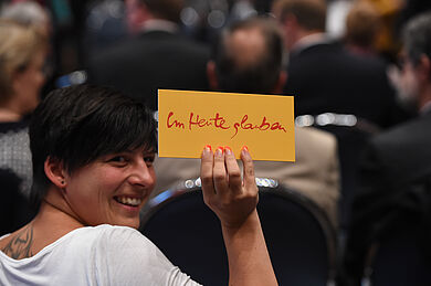 Eine junge Frau hält ein gelbes Schild mit dem roten Schriftzug "Im Heute glauben" hoch und lächelt.