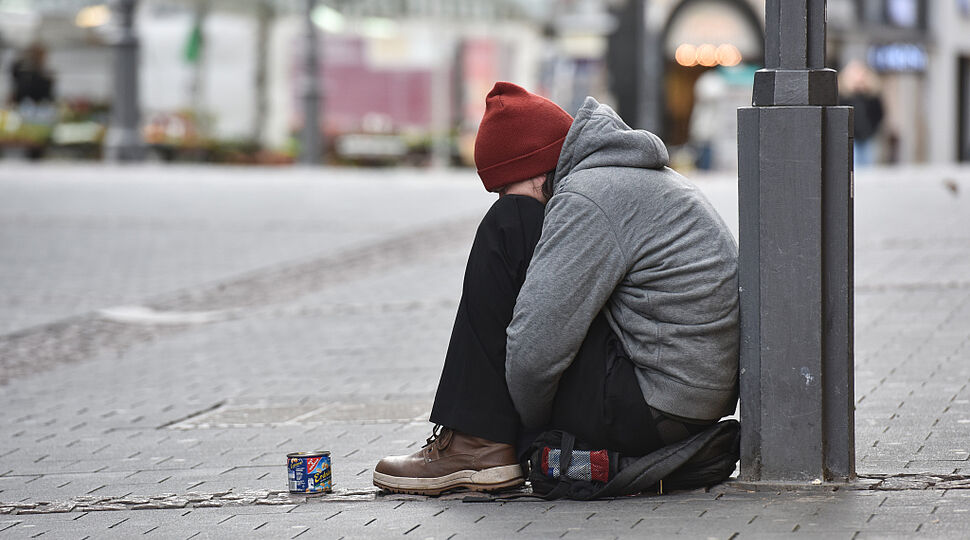 Ein Obdachloser sitzt, zusammengekauert auf dem Boden an einen Pfeiler gelehnt, in der Fußgängerzone. Vor ihm steht eine Blechdose. Sozialinitiative_170302-93-000141.jpg