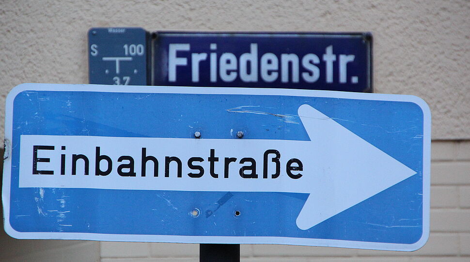 Ein Schild an der Mauer, auf dem Friedensstraße steht, davor ein Straßenschild mit der Aufschrift Einbahnstraße Bernhard_Riedl_1-bilder-erzbistum-koeln.de.JPG